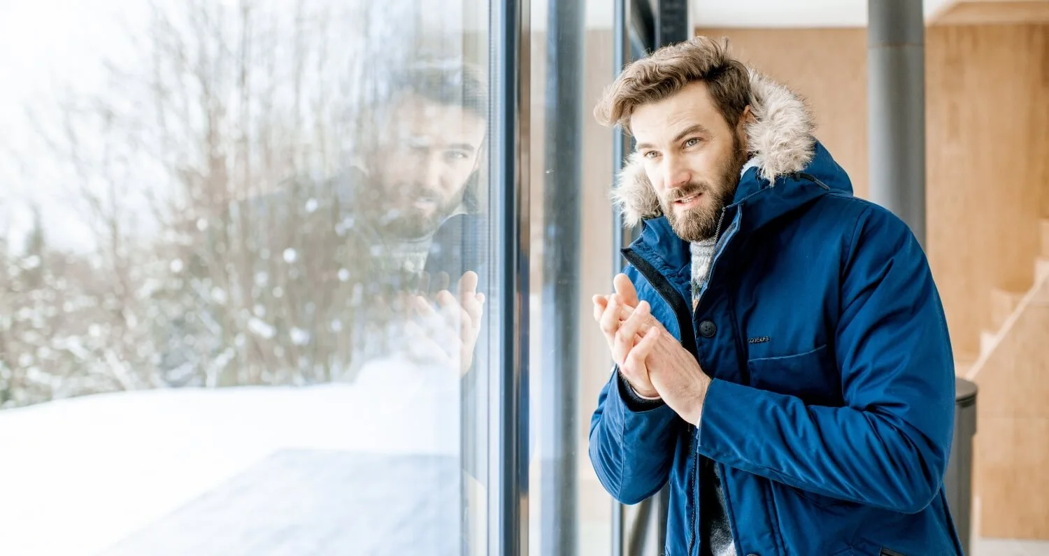 Man with a heavy winter jacket dresses inside the house while looking through the window.