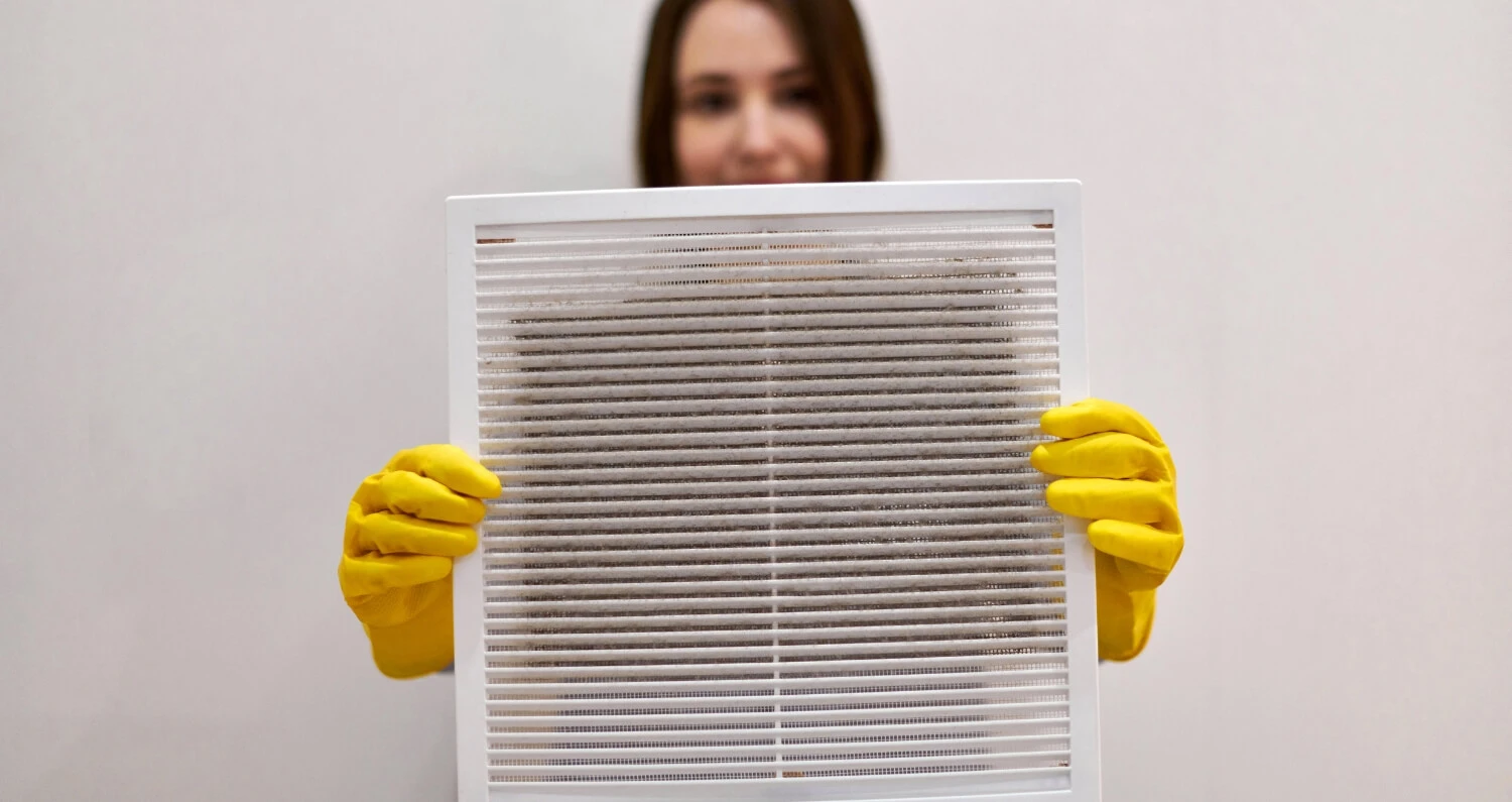 A woman with brown hair wears yellow gloves and is holding up a dirty vent with both hands.