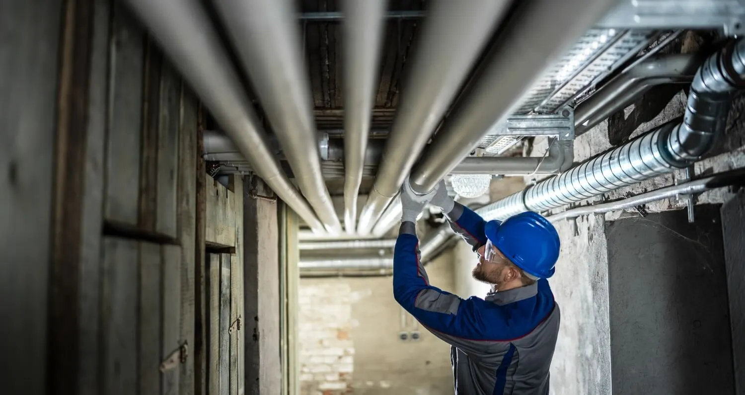 A male worker inspects pipes for leaks in a basement.